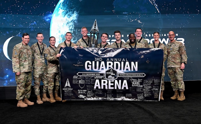 Finalists of the third annual Guardian Arena hold a banner to celebrate their accomplishments. Delta 9’s Team Einherjars, consisting of Capt. Coen Williams (holding a trophy), 1st Lt. Jakob Studtmann and 2nd Lt. Kaleb Holcomb (left to right of Williams), took home top honors in the competition, Dec. 8-9, that emphasized the importance of cultivating a combat-ready mindset, connecting military and civilian space enterprises, and building esprit de corps by putting the Guardian Ideals into action. Chief Master Sgt. of the Space Force John Bentivingna (far right) and Chief of Space Operations Gen. Chance Saltzman (far left) announced the top five teams following Bentivingna’s keynote address during the Space Force Association’s Spacepower Conference in Orlando, Fla., Dec. 11, 2025. The conference is an opportunity for U.S. Space Force senior leaders to meet and address Guardians, allies, partners and industry leaders. (U.S. Air Force photo by Chad Trujillo)