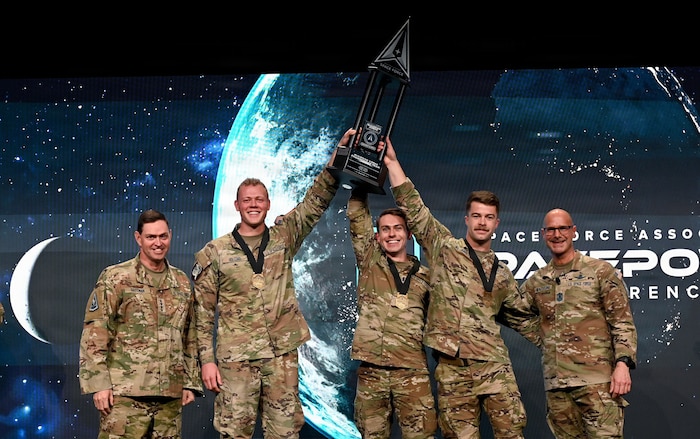 The winners of the third annual Guardian Arena – Delta 9’s Team Einherjars, consisting of Capt. Coen Williams, 1st Lt. Jakob Studtmann and 2nd Lt. Kaleb Holcomb (center left to right) – celebrate their victory as they hold a trophy to mark their accomplishment. Chief Master Sgt. of the Space Force John Bentivingna (far right) and Chief of Space Operations Gen. Chance Saltzman (far left) announced the top five teams following Bentivingna’s keynote address during the Space Force Association’s Spacepower Conference in Orlando, Fla., Dec. 11, 2025. The conference is an opportunity for U.S. Space Force senior leaders to meet and address Guardians, allies, partners and industry leaders. (U.S. Air Force photo by Chad Trujillo)