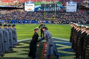 A midshipman wearing a Navy dress uniform and a soldier wearing a West Point dress uniform shake hands at midfield in a football stadium. They are surrounded by a line of midshipmen and cadets in similar attire to the left and right, who are standing at attention.