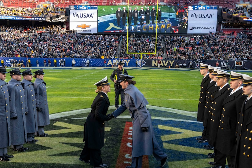 A midshipman wearing a Navy dress uniform and a soldier wearing a West Point dress uniform shake hands at midfield in a football stadium. They are surrounded by a line of midshipmen and cadets in similar attire to the left and right, who are standing at attention.