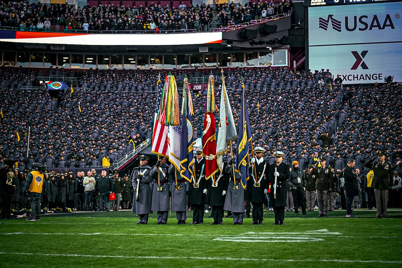 Eight people wearing military dress uniforms stand in a straight line in a stadium. Six of them are holding flags, including the American, Army and Navy flags, while the two standing to the far left and right of the line are holding rifles.