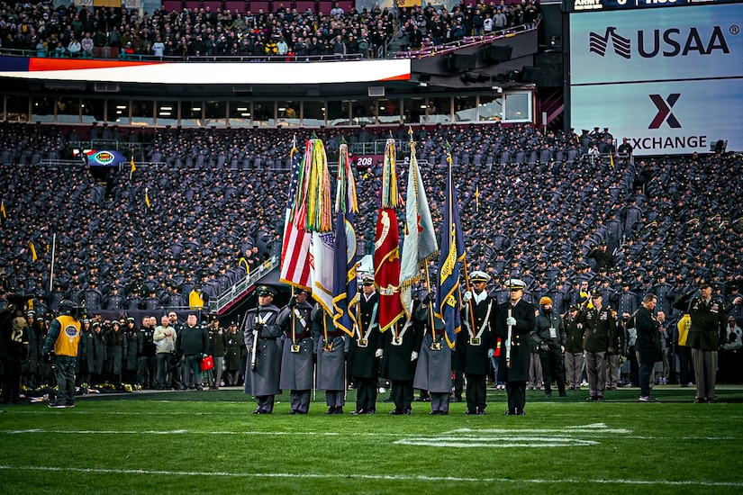 Eight people wearing military dress uniforms stand in a straight line in a stadium. Six of them are holding flags, including the American, Army and Navy flags, while the two standing to the far left and right of the line are holding rifles.