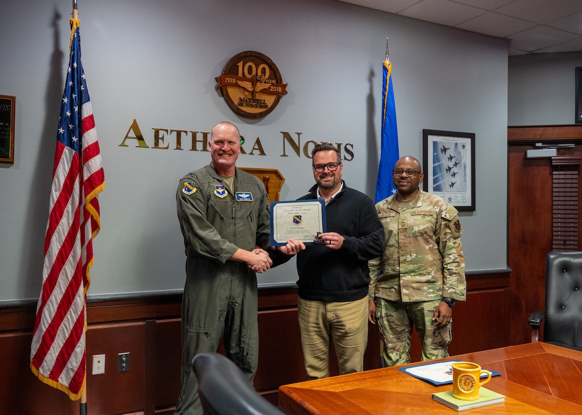 U.S Air Force Col. Adam Rudolphi, 42d Air Base Wing deputy commander, and Chief Master Sgt. Demetrius Booth, 42d ABW command chief, present the Crusader of the Month award for November to Ryan Hardeman, Maxwell Air Force base, Ala. Dec. 11, 2025.