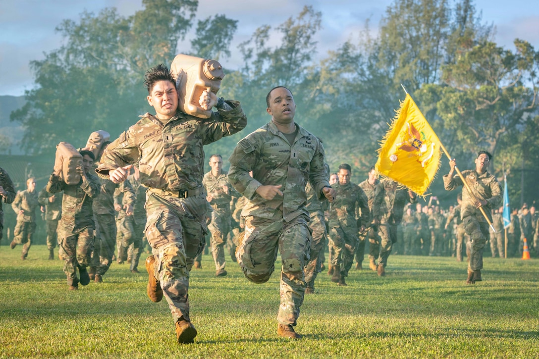 Soldiers, one carrying a flag and others carrying containers, run through a hazy field with trees in the background.