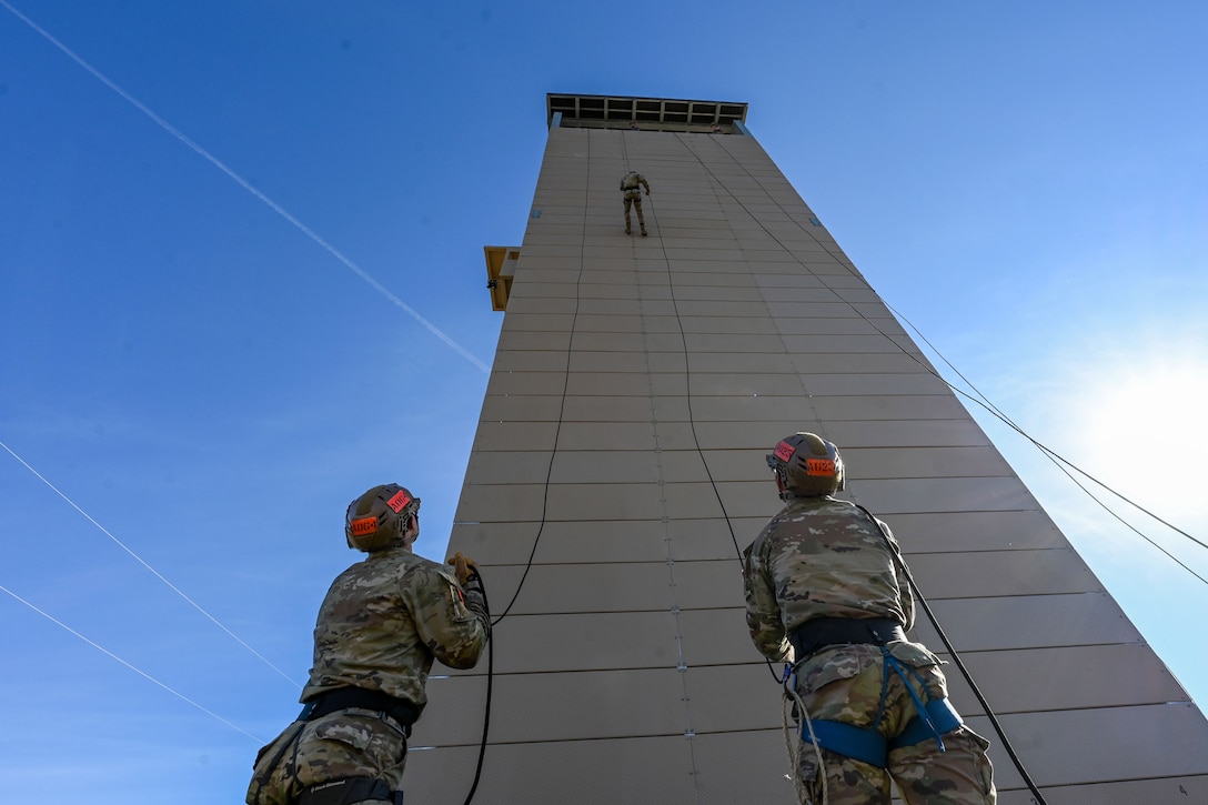 Two trainees hold rope while standing on the ground as a fellow trainee rappels down a large tower against a blue sky.