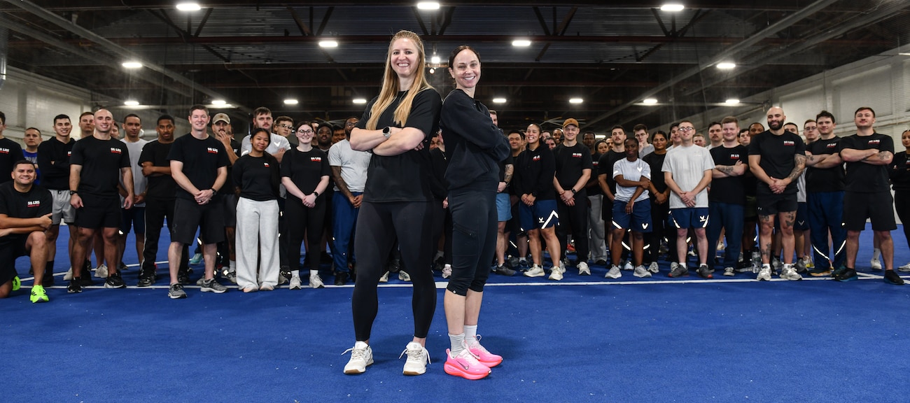 Kat Moake, physical training coach, and Lt. Col. Caitlin Oviatt, 55th Logistics Readiness Squadron commander, pose in front of the members of the 55th LRS during physical training at the Offutt Field House, December 12, 2025. Moake was hired to lead a more strenuous version of PT to ensure LRS members exceed PT requirements. (U.S. Air Force photo by Chad Watkins)