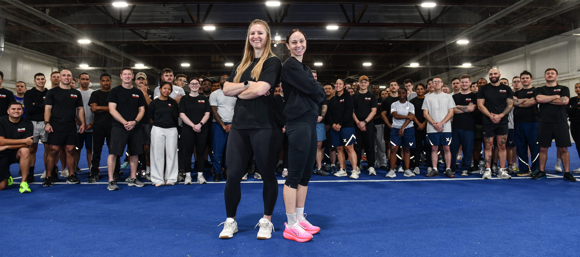 Kat Moake, physical training coach, and Lt. Col. Caitlin Oviatt, 55th Logistics Readiness Squadron commander, pose in front of the members of the 55th LRS during physical training at the Offutt Field House, December 12, 2025. Moake was hired to lead a more strenuous version of PT to ensure LRS members exceed PT requirements. (U.S. Air Force photo by Chad Watkins)