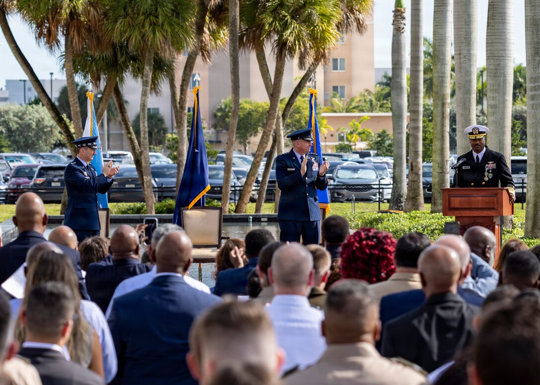 A man in a black military unform address a large crowd from behind a podium.