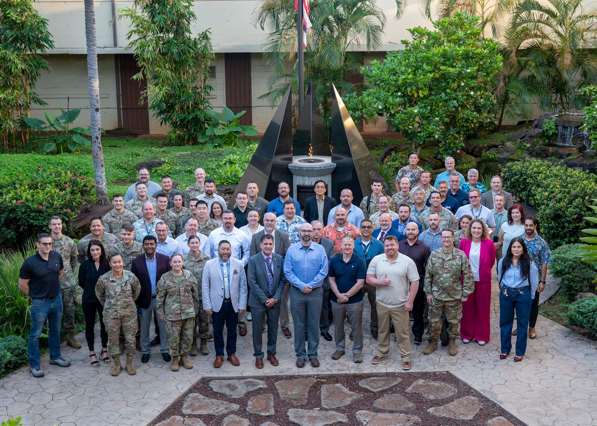 A group of civilians dressed in professional attire and service members in the Air Force Airman Combat Uniform, resembling a camouflage pattern, from Pacific Air Forces directorates and DAF organizations pose for a group photo in front of the memorial in the Courtyard of Heroes where an eternal flame is lit and flickering behind them.
