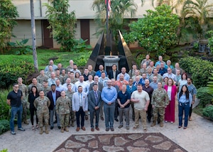 A group of civilians dressed in professional attire and service members in the Air Force Airman Combat Uniform, resembling a camouflage pattern, from Pacific Air Forces directorates and DAF organizations pose for a group photo in front of the memorial in the Courtyard of Heroes where an eternal flame is lit and flickering behind them.