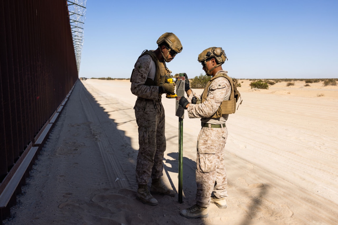 A man in a camouflage military uniform uses a drill to attach a warning sign to a pole, while another man in similar attire holds the sign steady. Both men are standing in a desert location with a long fence next to them.