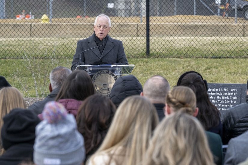 A man in cold-weather attire stands at a podium outside, speaking to a crowd.