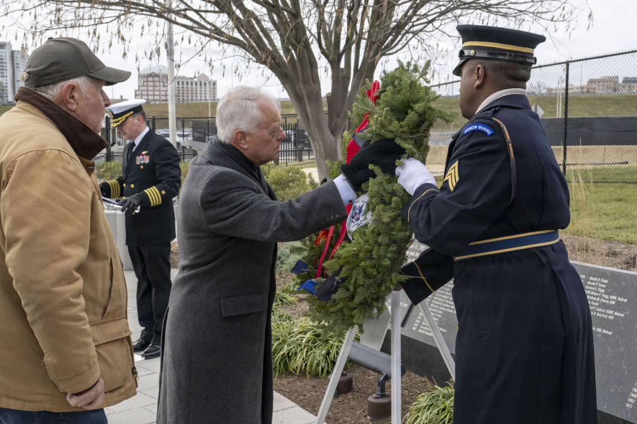 A man in cold-weather attire and another man in a military dress uniform place a wreath on an easel at a memorial outside, as another man in cold-weather attire looks on.