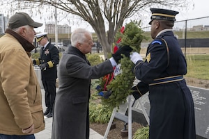 A man in cold-weather attire and another man in a military dress uniform place a wreath on an easel at a memorial outside, as another man in cold-weather attire looks on.