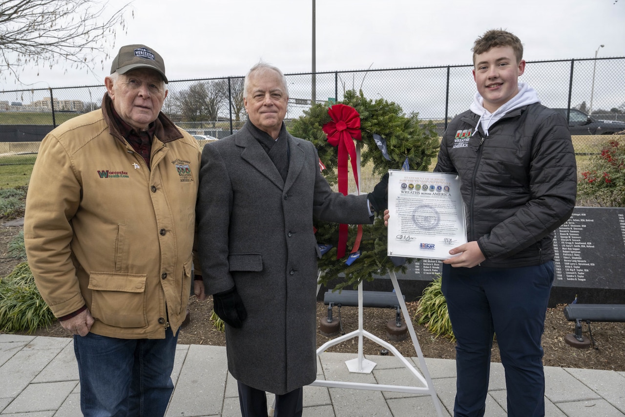 Three men in cold-weather attire stand outside in front of a wreath.