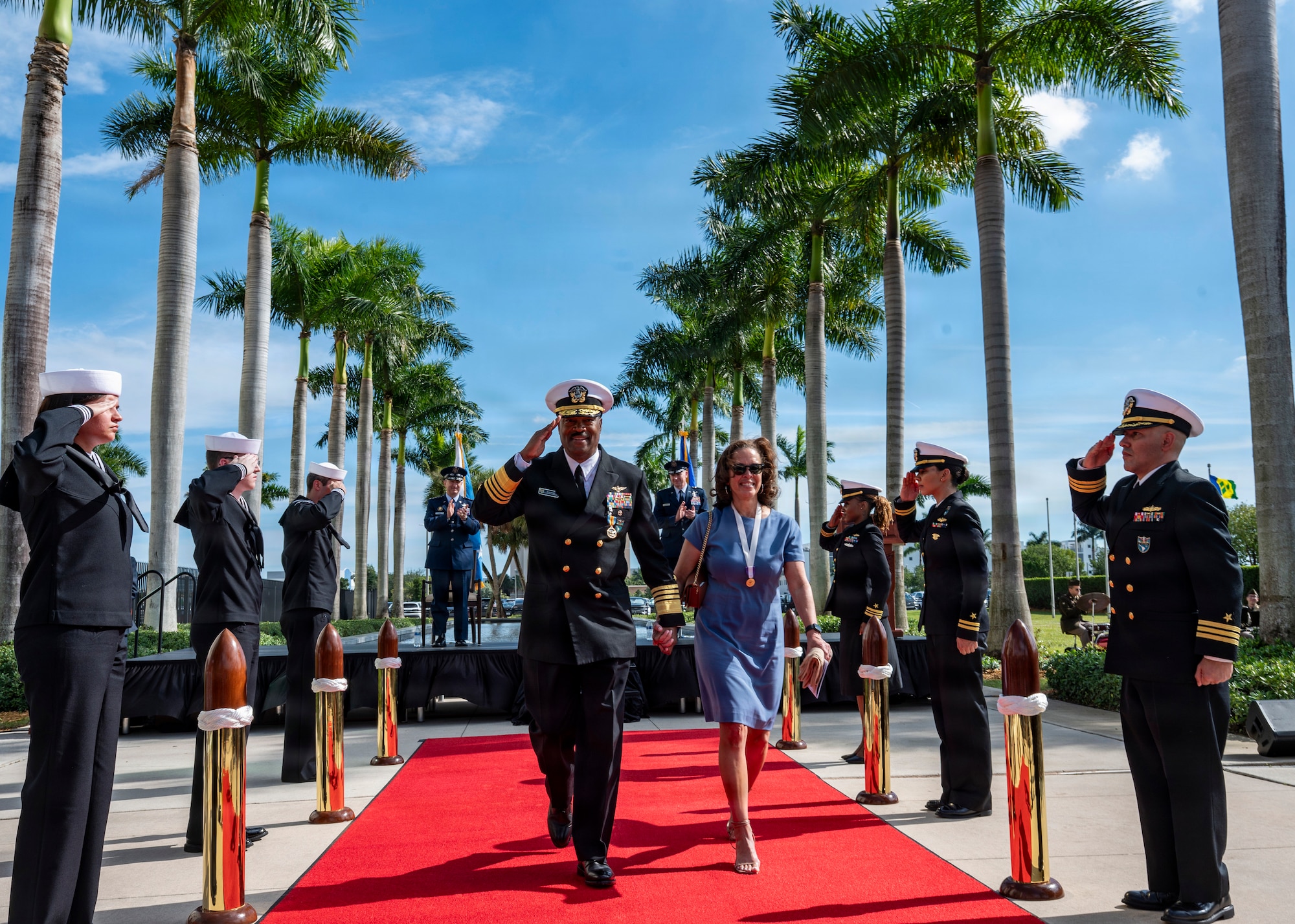 A man in a black military unform and a woman in a blue dress walk a red carpet while flanked by uniform personnel saluting.