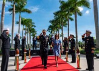 A man in a black military unform and a woman in a blue dress walk a red carpet while flanked by uniform personnel saluting.