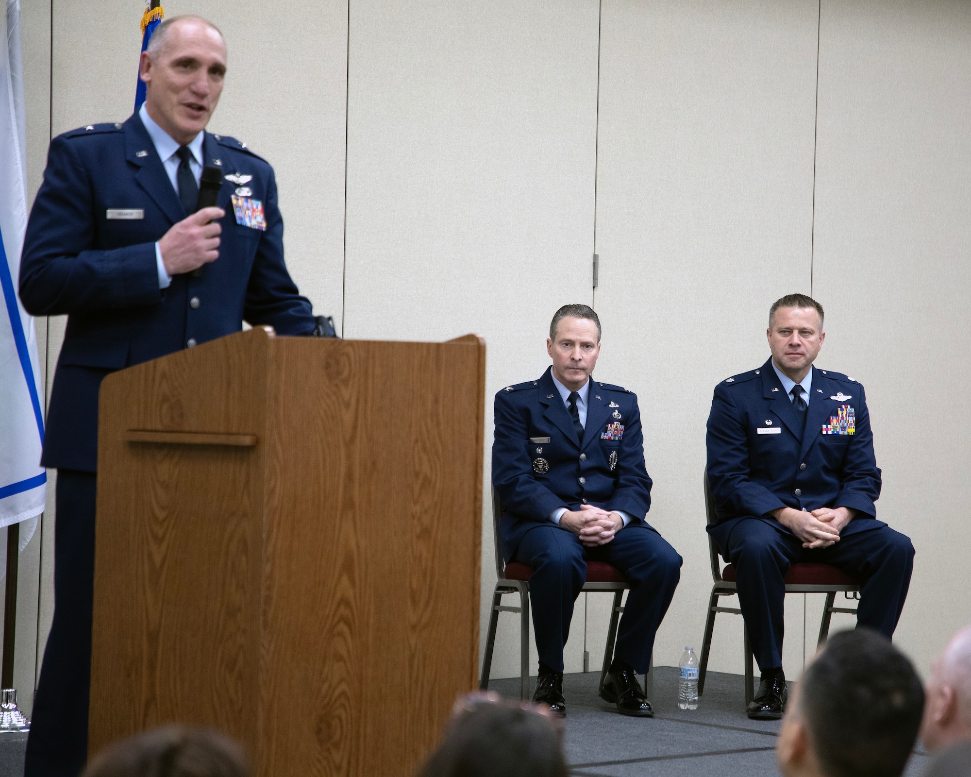 Male Air Force officer speaks at podium while two other officers sit to his left on stage.