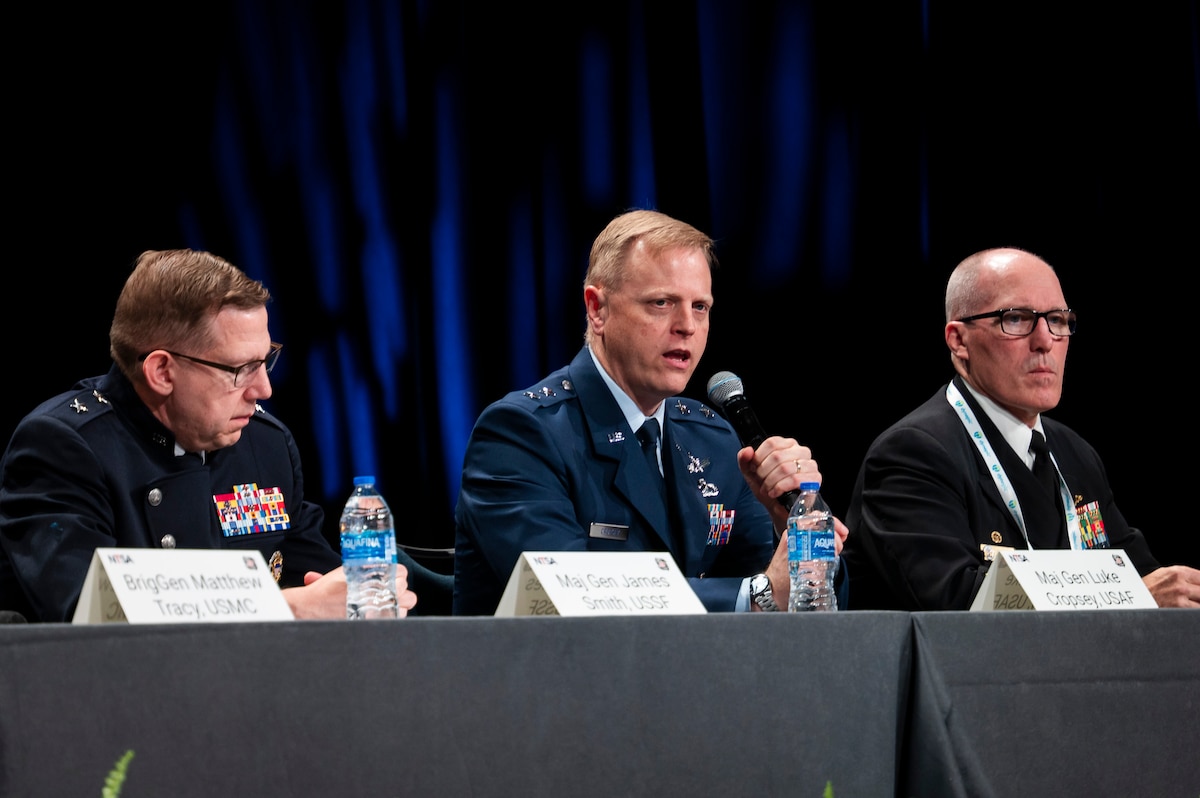 A Space Force and Air Force general are joined on stage by a man in a black business suit. Holding mics and talking to crowd. Each man has a table tent with his name.