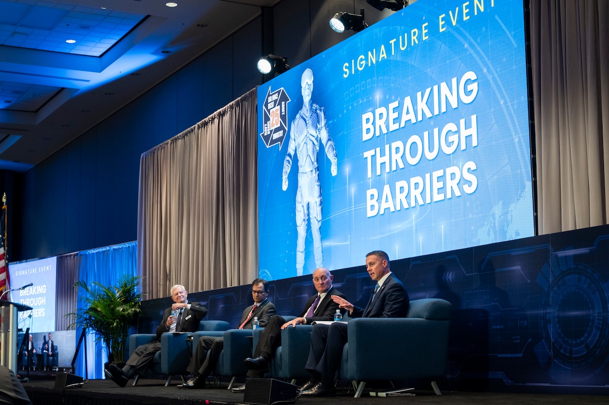 Four men sit on stage in suits. Banner behind them says Breaking Through Barriers.