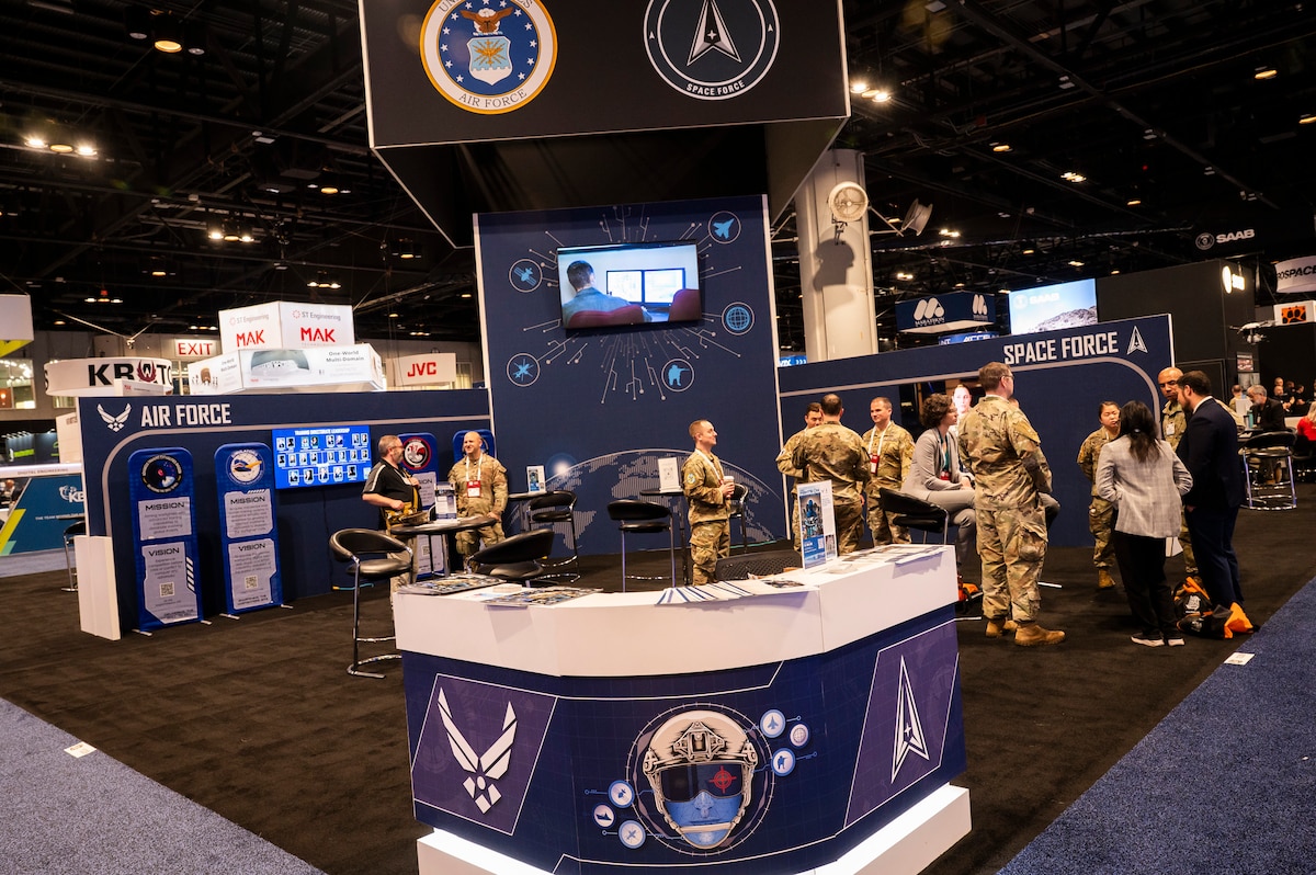 Individuals in Air Force uniforms stand in conference booth that features Air Force and Space Force logos and blue carpet.