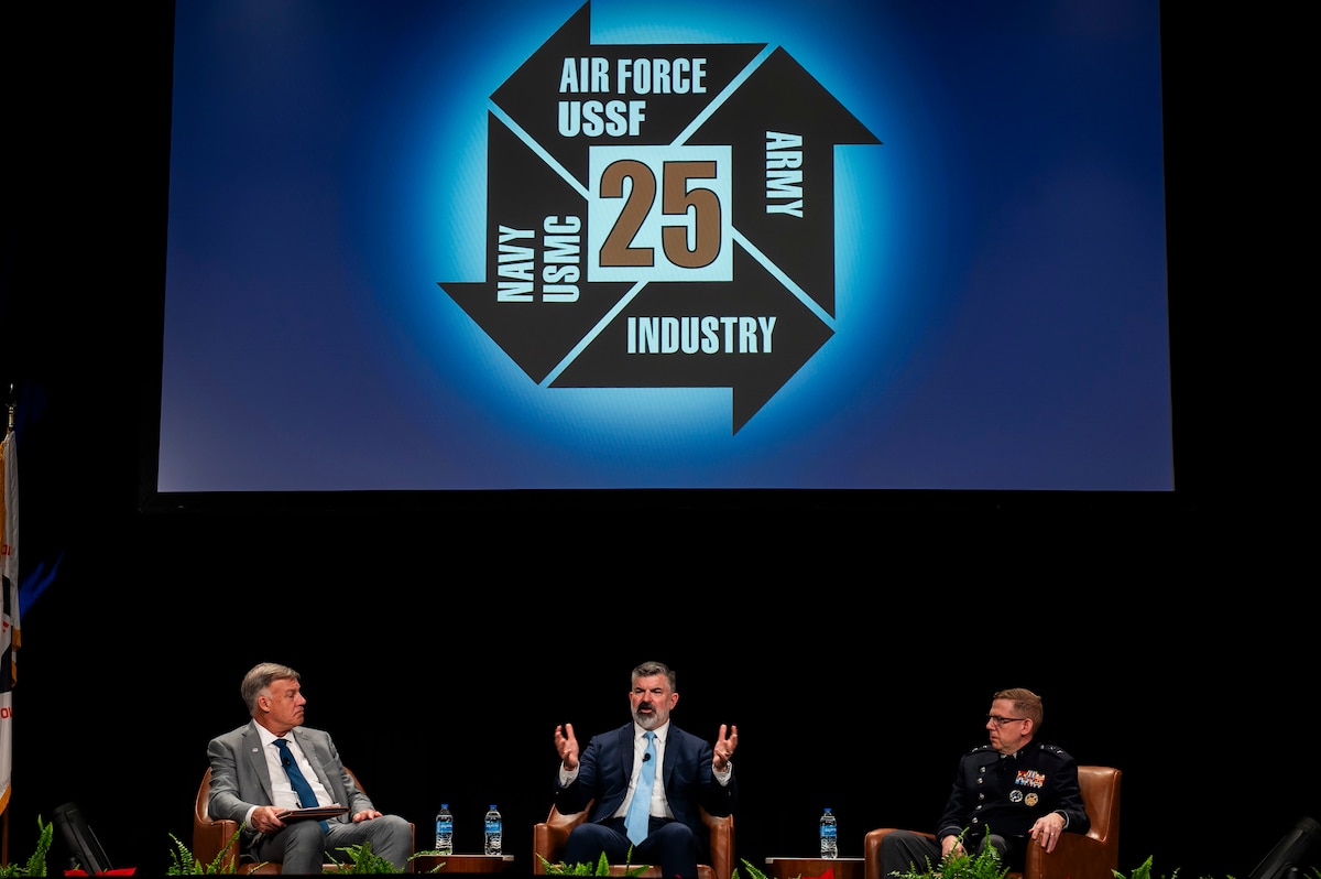 Men in suits sit in chairs on dark stage. Banner has the I/ITSEC conference logo in blue.