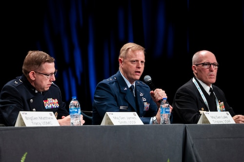 A Space Force and Air Force general are joined on stage by a man in a black business suit. Holding mics and talking to crowd. Each man has a table tent with his name.