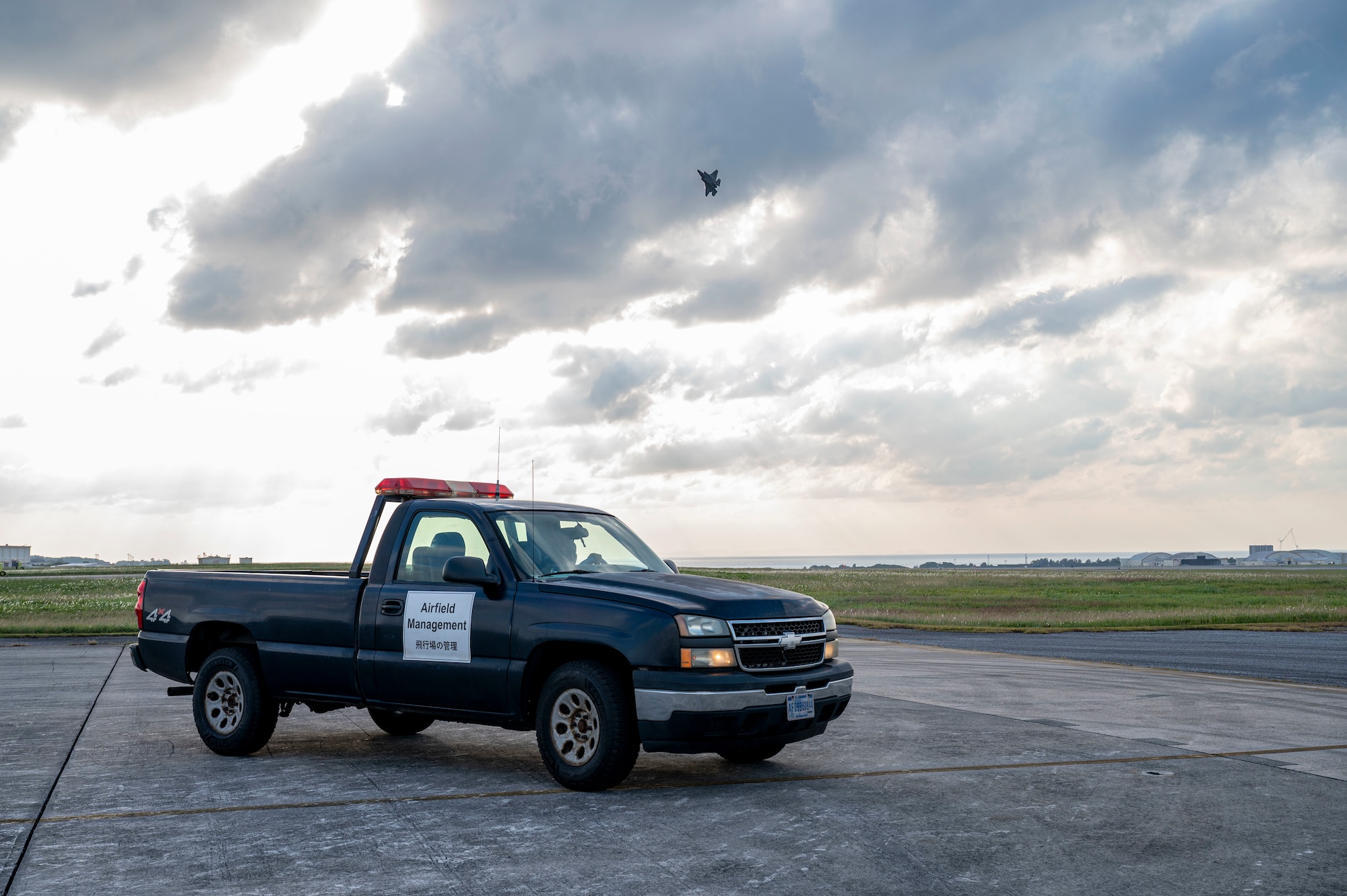 An 18th Operations Support Squadron airfield management vehicle drives on the flight line as a U.S. Air Force F-35 Lightning II assigned to the 356th Expeditionary Fighter Squadron, deployed to Kadena Air Base, flies overhead at Kadena Air Base, Japan, Dec. 11, 2025.