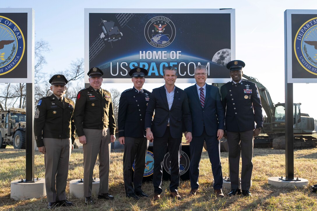 The secretary of war alongside military leaders pose for a photo in front of a sign in a field.
