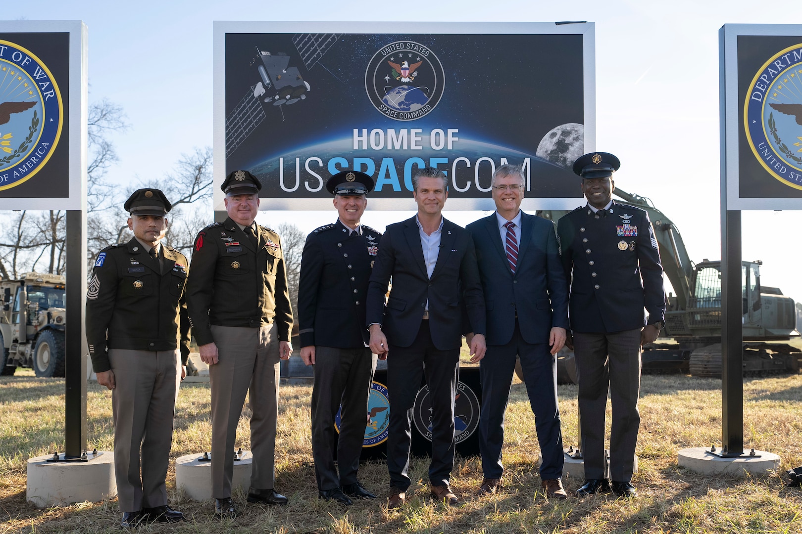 The secretary of war alongside military leaders pose for a photo in front of a sign in a field.