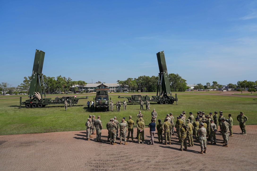 Service members and civilians stand in a large field near two weapon systems mounted on trailers flanking a parked military vehicle.