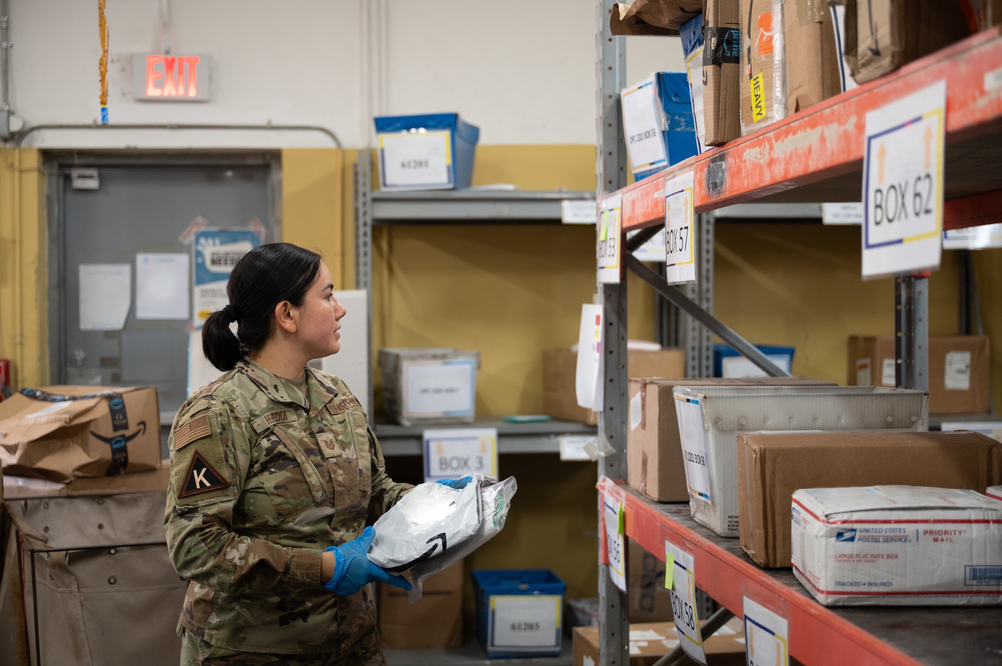 An Airman holding a package stands to the left of a rack filled with packages and mail bins
