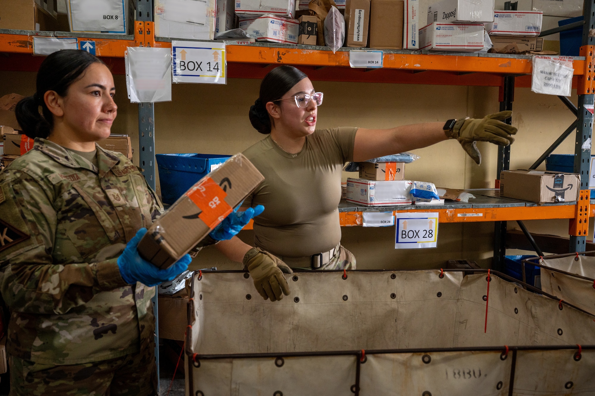 An Airman holds a box over a container while another Airman points to the right of the frame while speaking