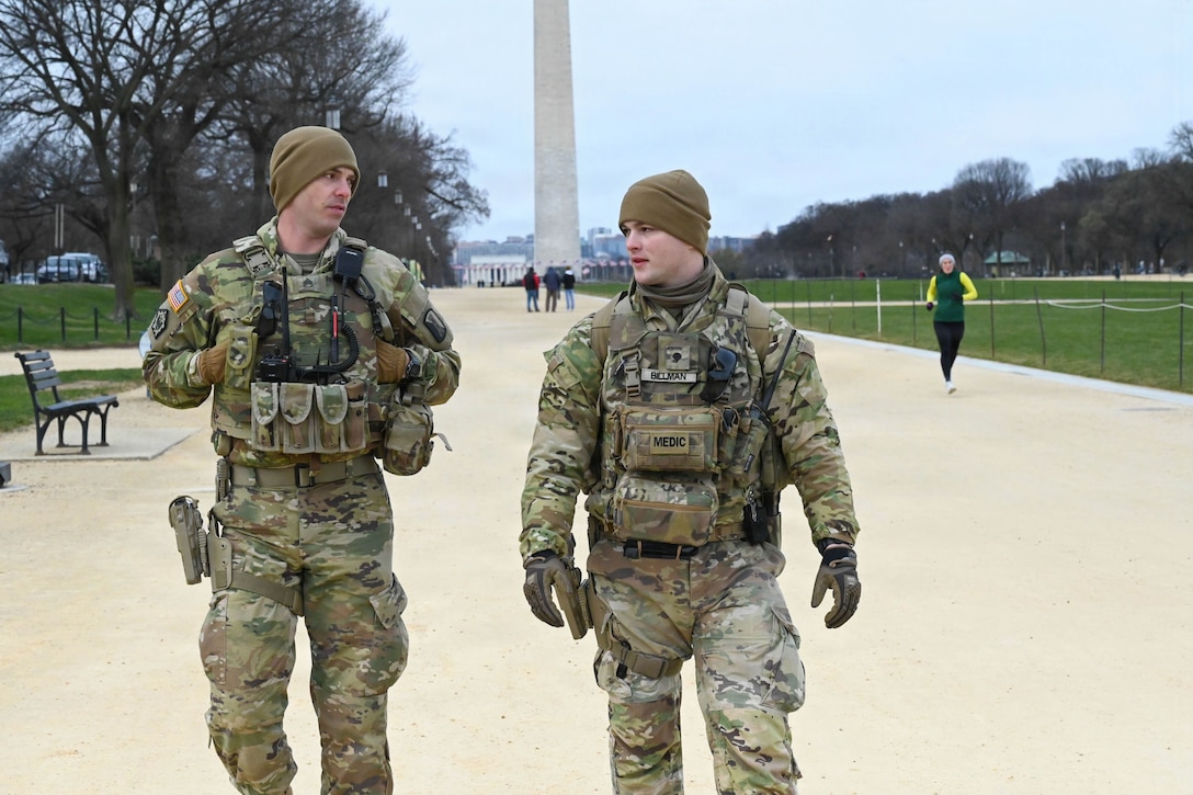 Two soldiers walk on a dirt path in front of a tall monument on a gloomy day.