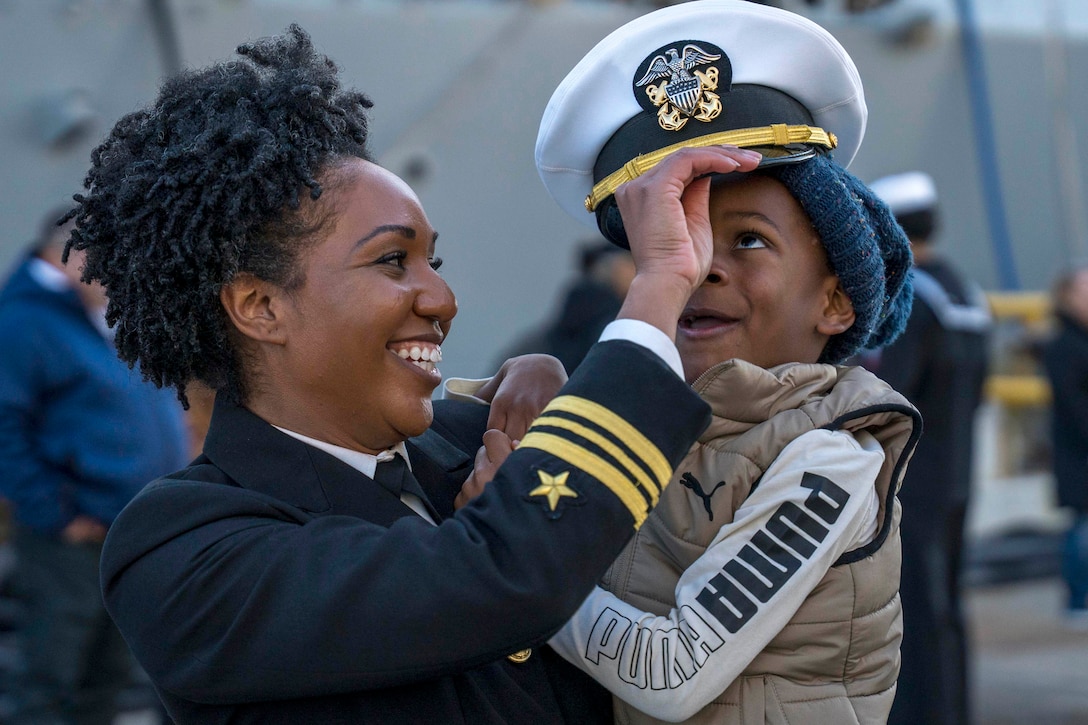 A smiling sailor places their hat on child they're holding in front of a blurred partially visible ship in the background.