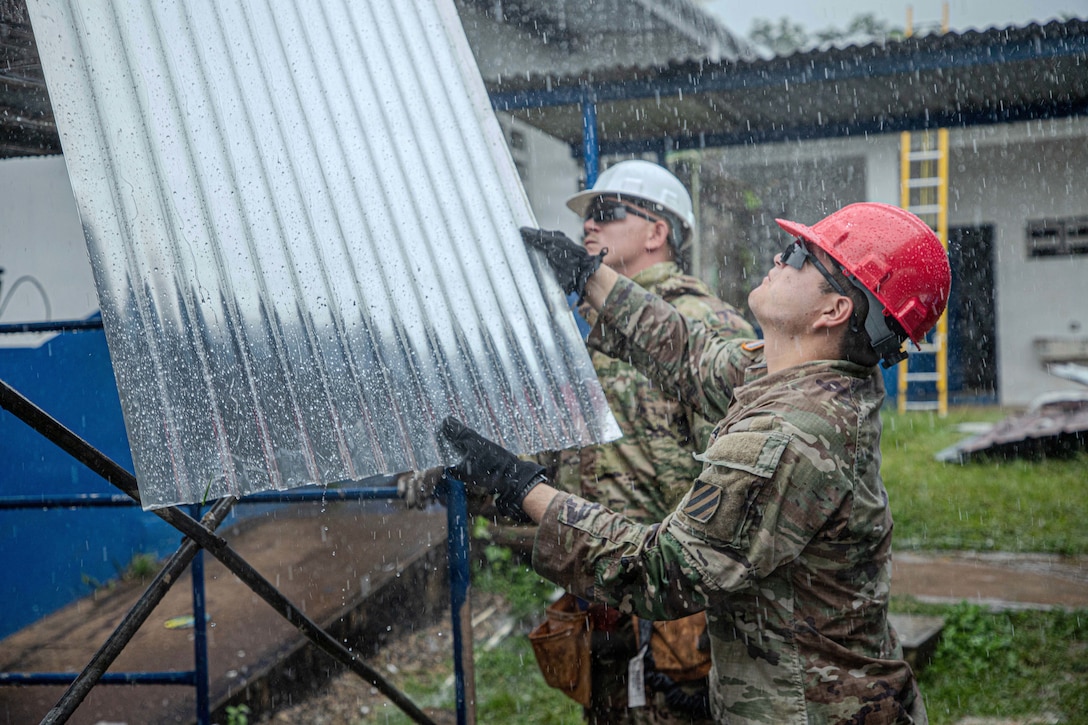 Two soldiers in hard hats, one holding a panel, repair a school roof in the rain.