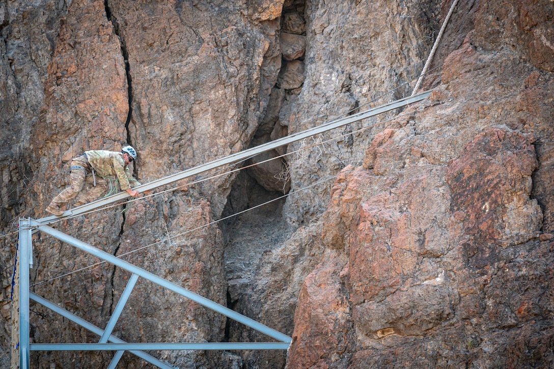 A service member wearing a hard hat and harness climbs a metal structure attached to a mountain.