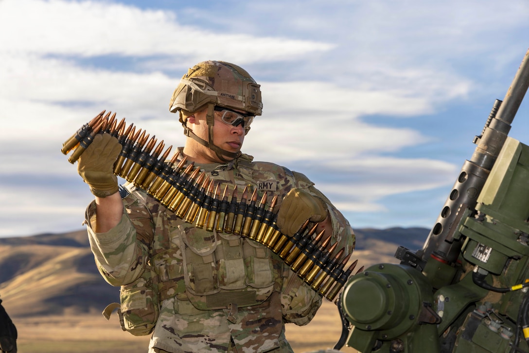 A soldier in tactical gear loads belts of ammo into a weapon in a hilly area under a blue, cloudy sky.