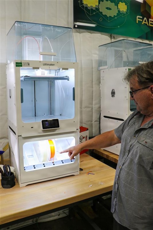 A man wearing casual attire, standing to the right, points at a 3D printer.