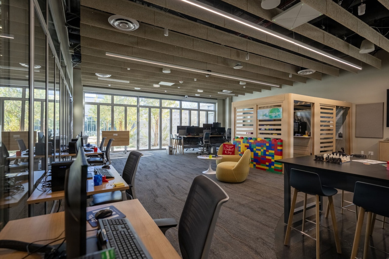 The interior of an office space is shown with desks, computers and chairs. There is a wall of windows at the far end of the room.