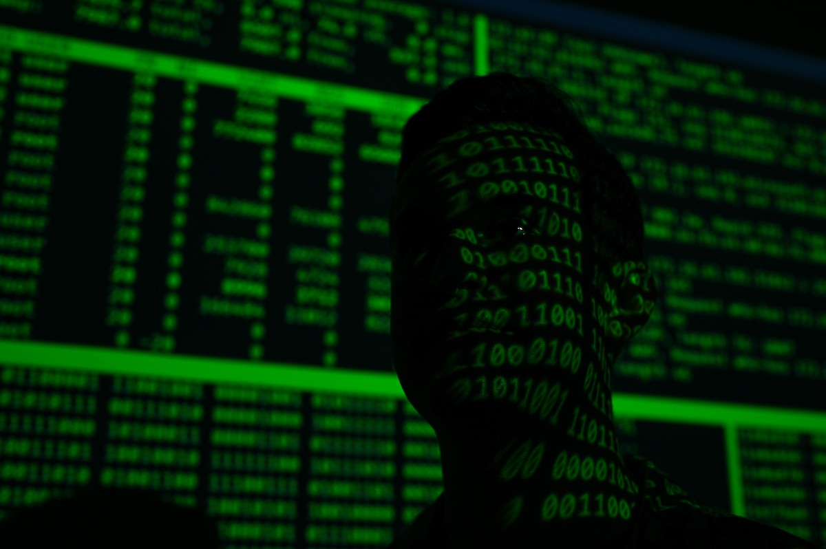 A silhouetted head is seen with green binary code displayed over their face, with green and black code displayed behind them.
