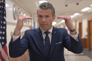 A man in a blue suit holds up his hands level while walking down a hallway indoors next to a flag.