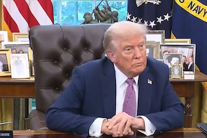 President Trump is sitting at a desk in the Oval Office with standing flags behind.