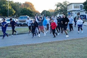 People in different colored athletic attire run on the curve of a street away from a parking lot.