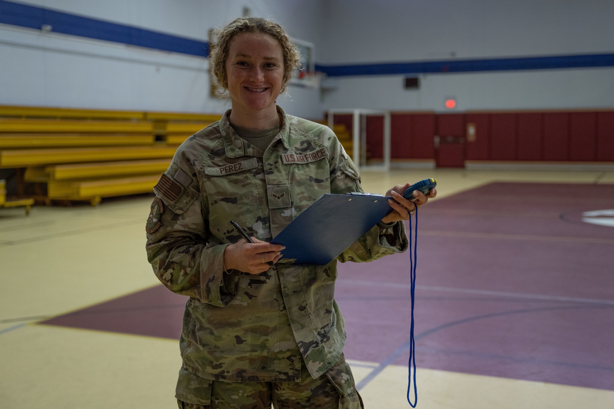 Airman 1st Class Jasmine Perez stands in uniform in a gymnasium holding a clipboard and stop watch smiling at the towards the camera.