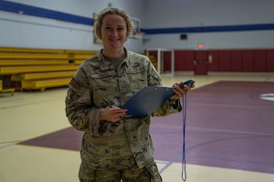 Airman 1st Class Jasmine Perez stands in uniform in a gymnasium holding a clipboard and stop watch smiling at the towards the camera.