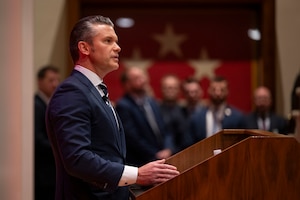 A side view of the secretary of war speaking at a podium with others watching in the blurred background.