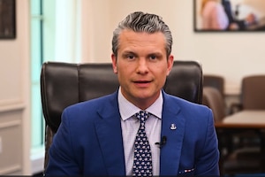 The secretary of war sits at a desk while speaking to a camera in an office.