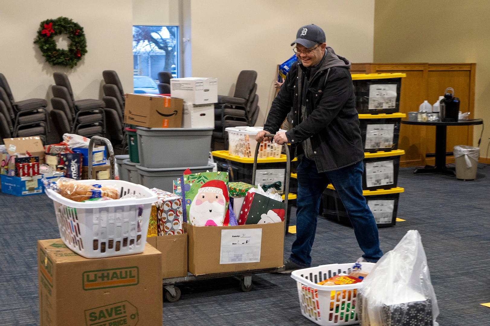 A man in a heavy winter coat pushes a flat bed cart with packages on it through a room full of Christmas packages