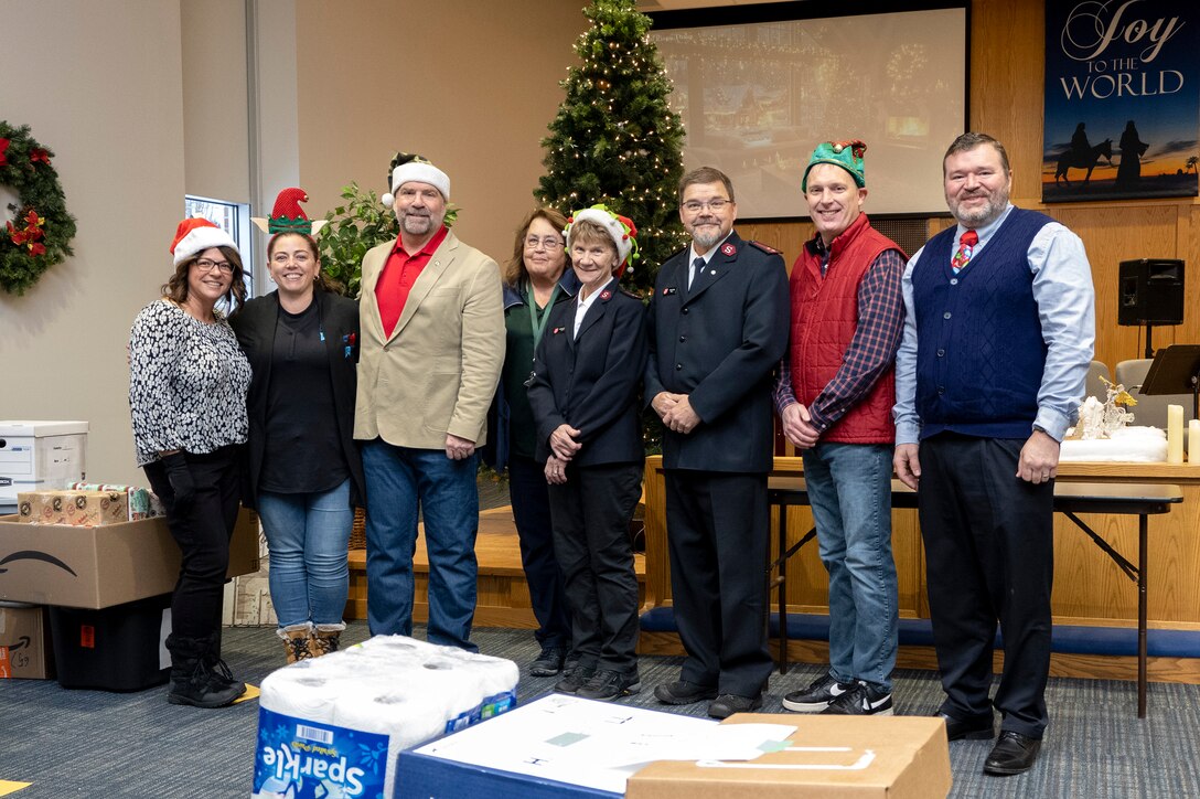 eight people stand in a room with a Christmas tree in the background and donated items around them.